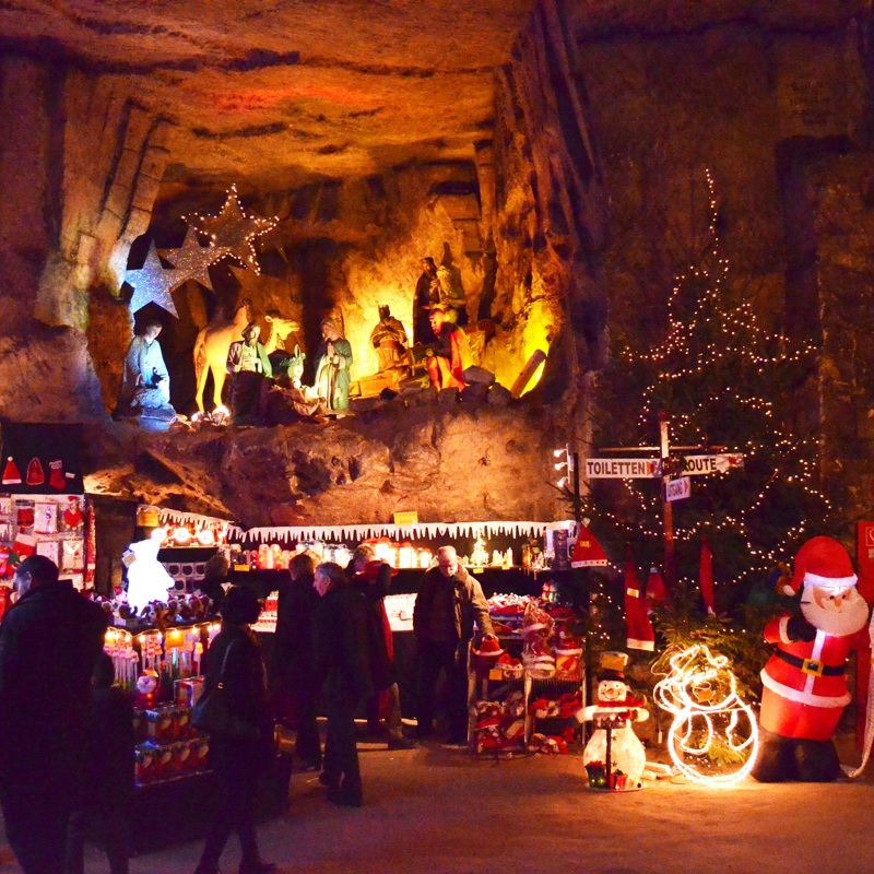 Shoppende mensen bij verschillende kerststands in de gemeentegrot. Een extra gang op hoogte waarin een grote kerststal is gepositioneerd en uitgelicht.